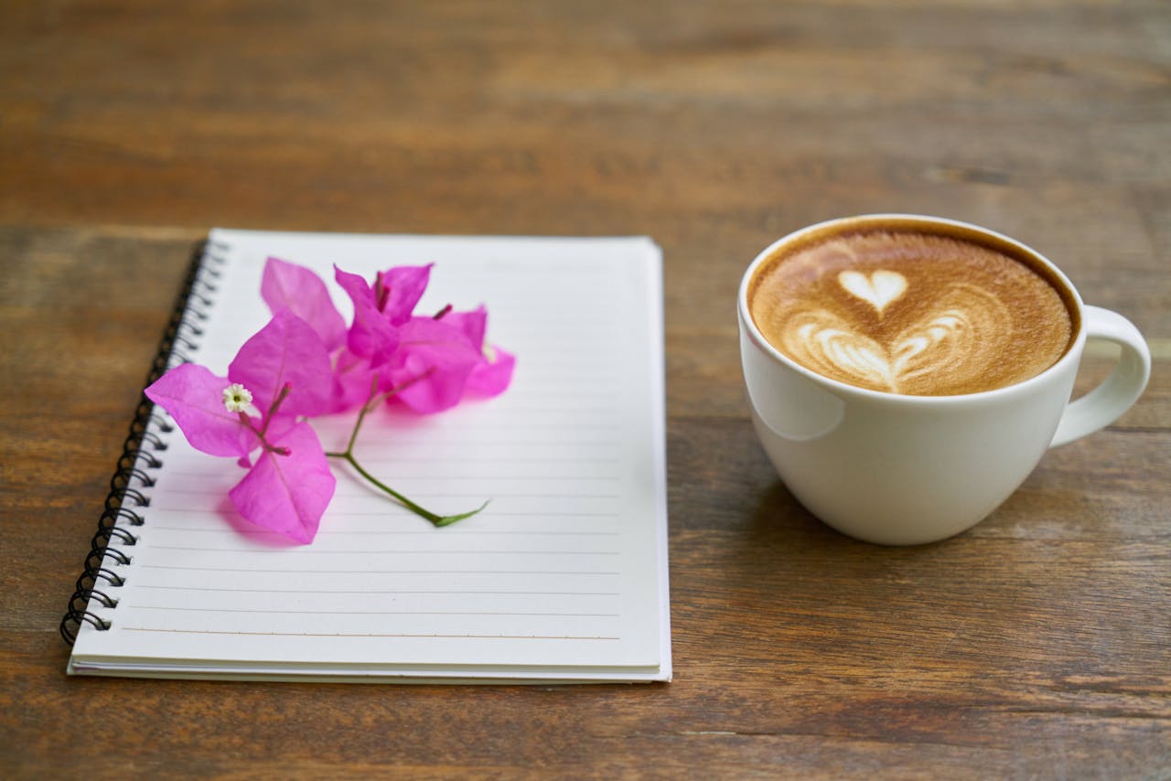 A close-up of a cappuccino with latte art and a notebook adorned with pink flowers on a wooden table.