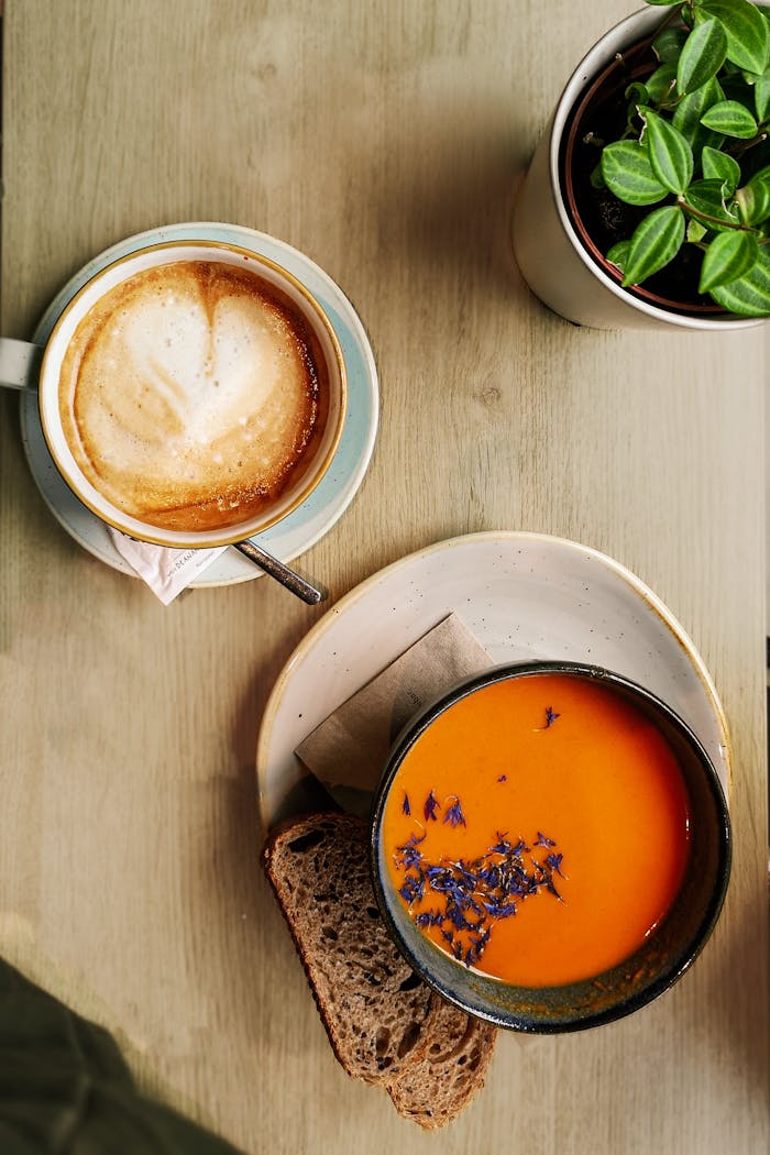 Close-up view of cappuccino, tomato soup garnished with flowers, and bread on a wooden table.