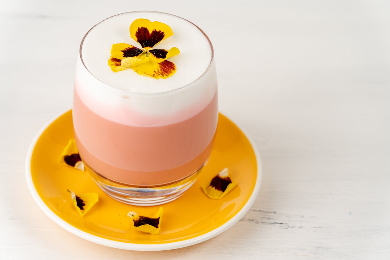 A vibrant floral latte topped with colorful edible pansies, presented in a glass on a bright yellow saucer.