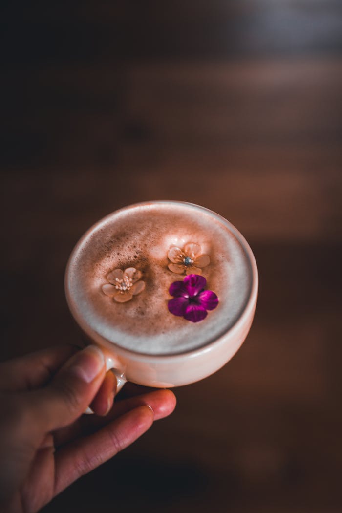 A close-up of a coffee cup with artistic foam and flowers, held in hand.